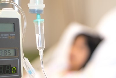 A closeup of an IV drip with a patient lying on a bed in the background out-of-focus.