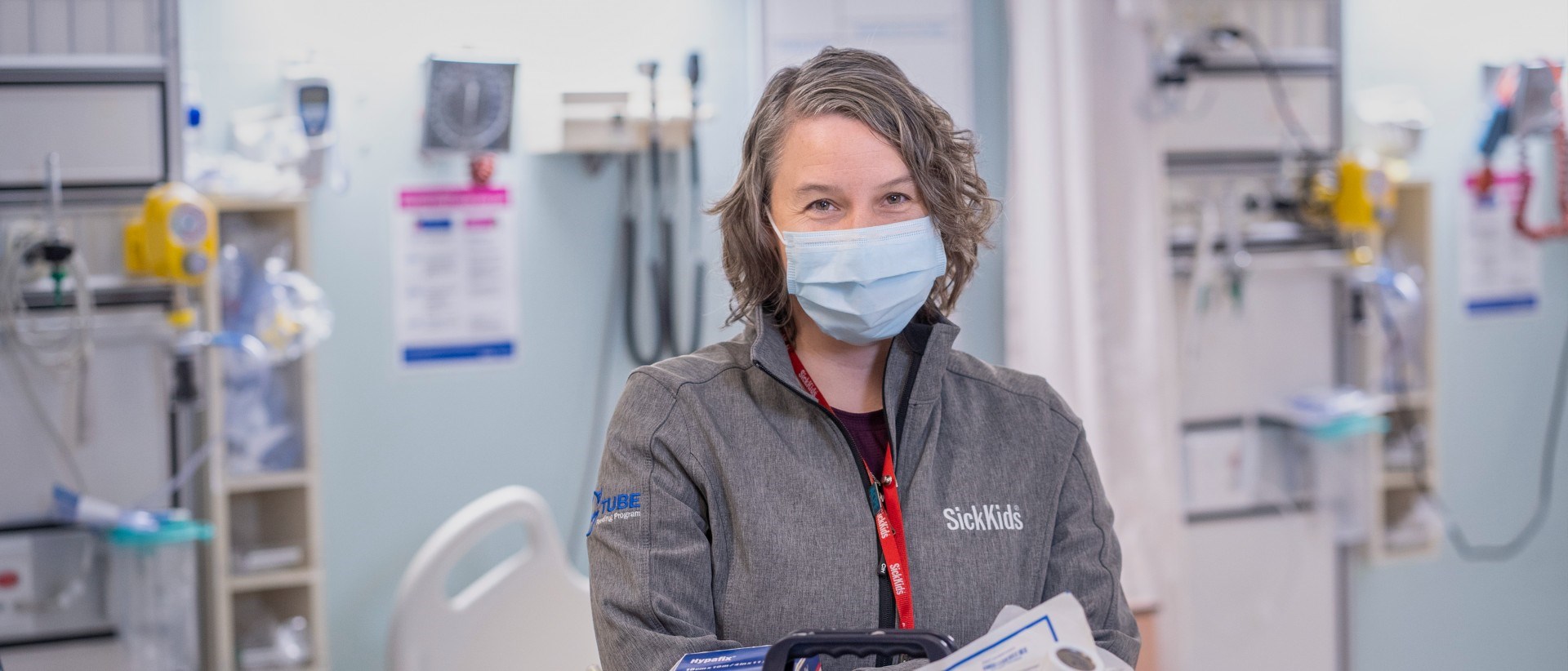 Holly Norgrove wearing a mask in a hospital room equipped with various devices and clinical equipment. She is wearing a sweater with stitching that says "SickKids" and "G-Tube feeding program".