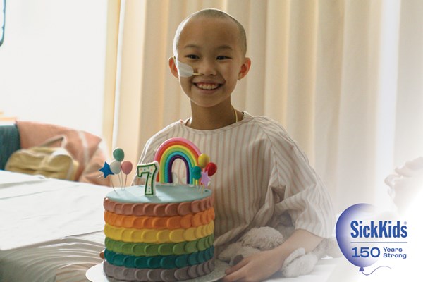 A smiling child sitting on a hospital bed and holding a rainbow birthday cake with a "7" candle on top. SickKids 150 Years Strong.