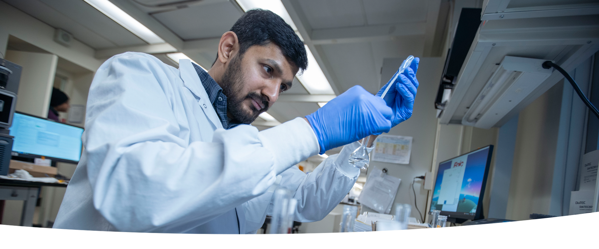 A SickKids staff member wearing a lab coat and blue gloves carefully pours a liquid from a pipette into a flask in a laboratory setting. He is focused on his task, with lab equipment and a computer screen visible in the background.