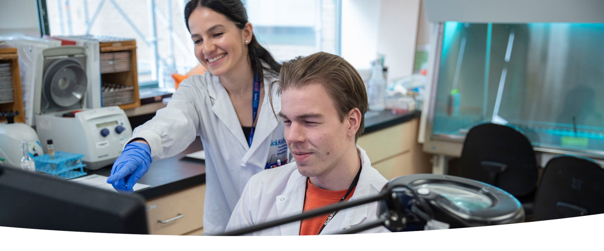 Two lab professionals at a lab bench. One is seated in front of a computer screen while the other is standing over and pointing at the screen.