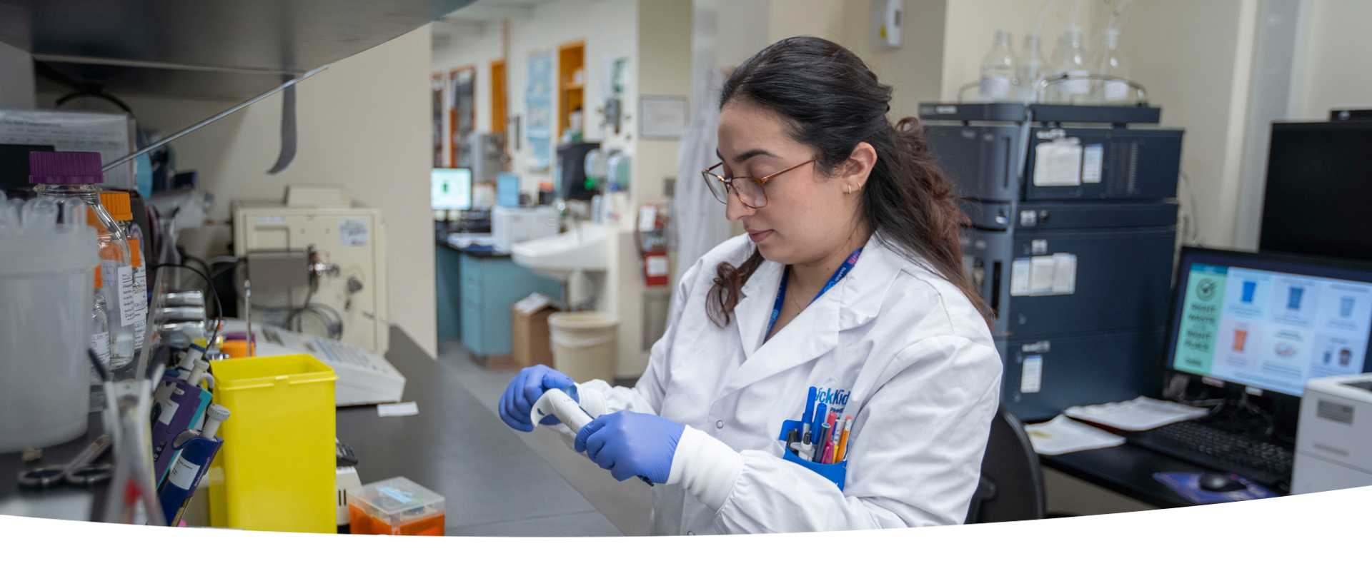 A lab professional wearing a lab coat and blue gloves. She is handling a piece of equipment at a lab bench.