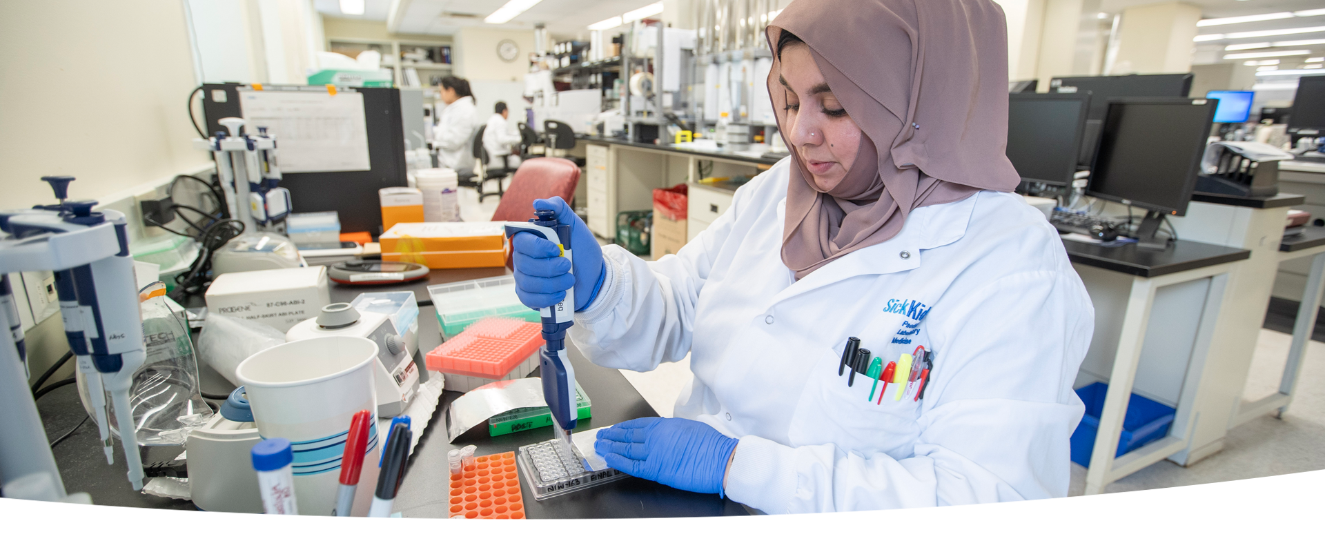 A laboratory professional pipetting into a multiwell plate at a lab bench. In the background, there are other lab professionals at work.