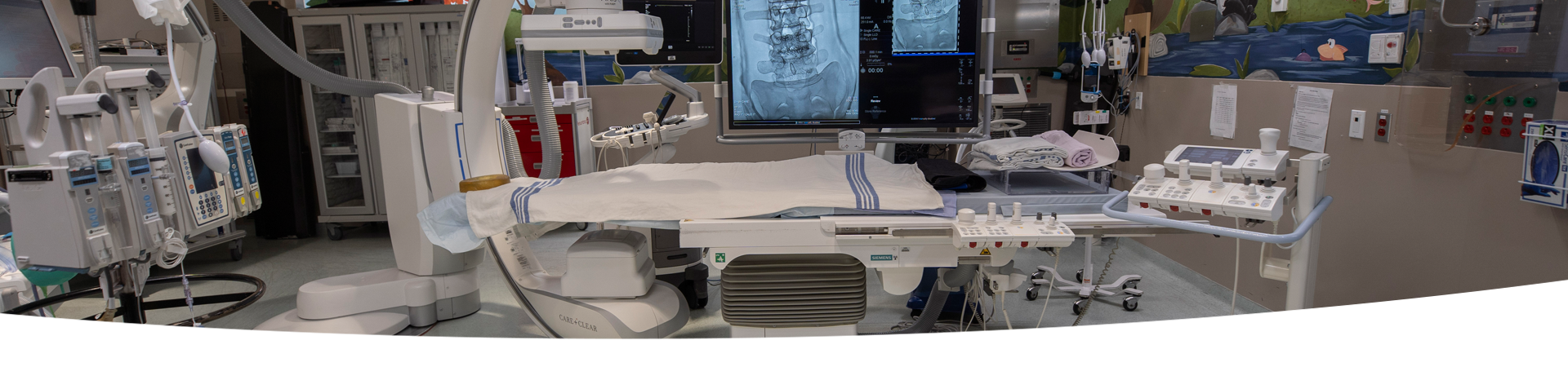 A room with a Single Plane Digital Angiography unit. There is a variety of hospital equipment in the background.