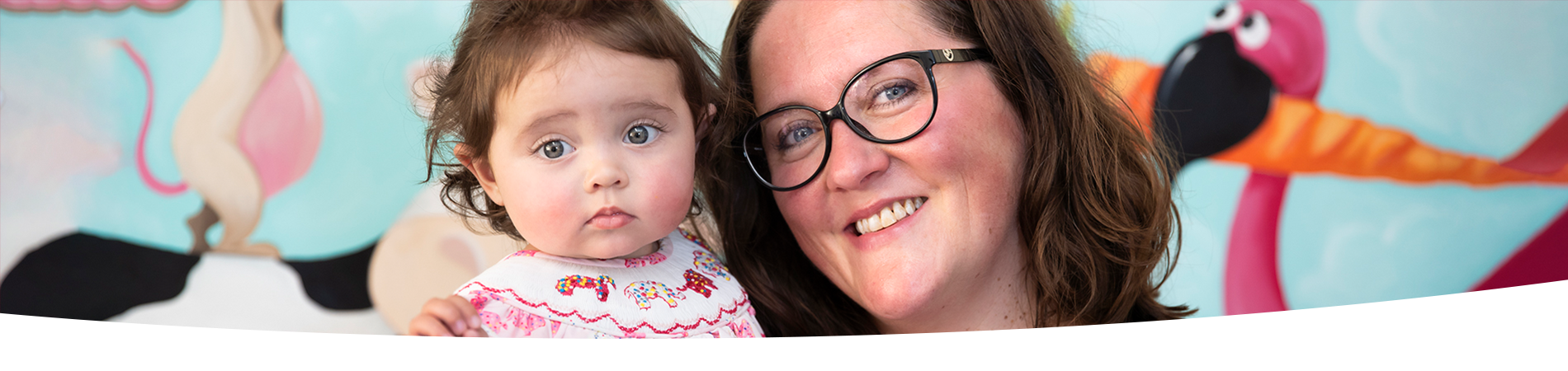 An infant and a woman in glasses pose together for a photo in front of a colourful mural. The mural has paintings of cartoon animals, including a cow, flamingo, and mouse.