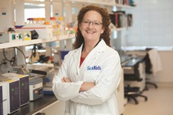 Woman stands in a lab wearing a lab coat.