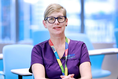 Nurse Jane Stuart-Minaret sits in a classroom with blue chairs and desks with a window in the background, wearing glasses, a SickKids lanyard and a purple shirt.