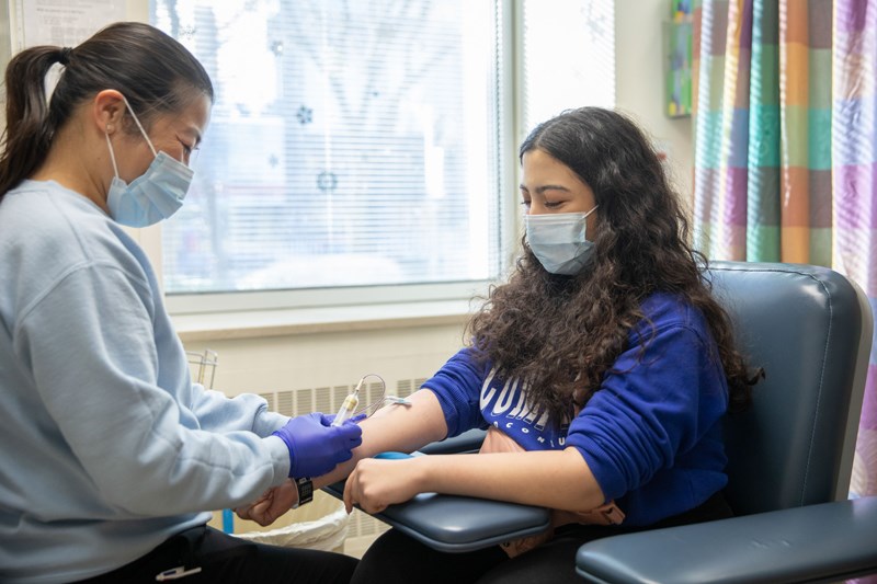 Melika seated in a chair with her sleeves rolled up while getting her blood drawn by a phlebotomist.