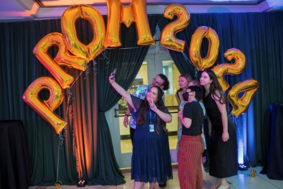 A SickKids staff member taking a group selfie with SickKids 2024 prom-goers. There are golden balloons spelling out Prom 2024 in the background.