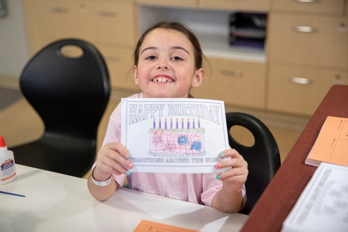 A child smiling and holding up a colouring page of a birthday cake.