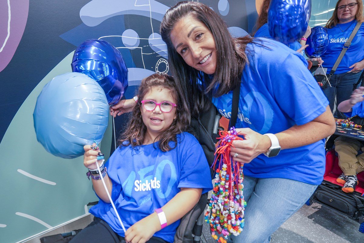 Morgan and Chloe posing for a photo. Both are dressed in blue SickKids 150 t-shirts and holding blue balloons. Morgan is carrying Bravery Beads in her other hand.