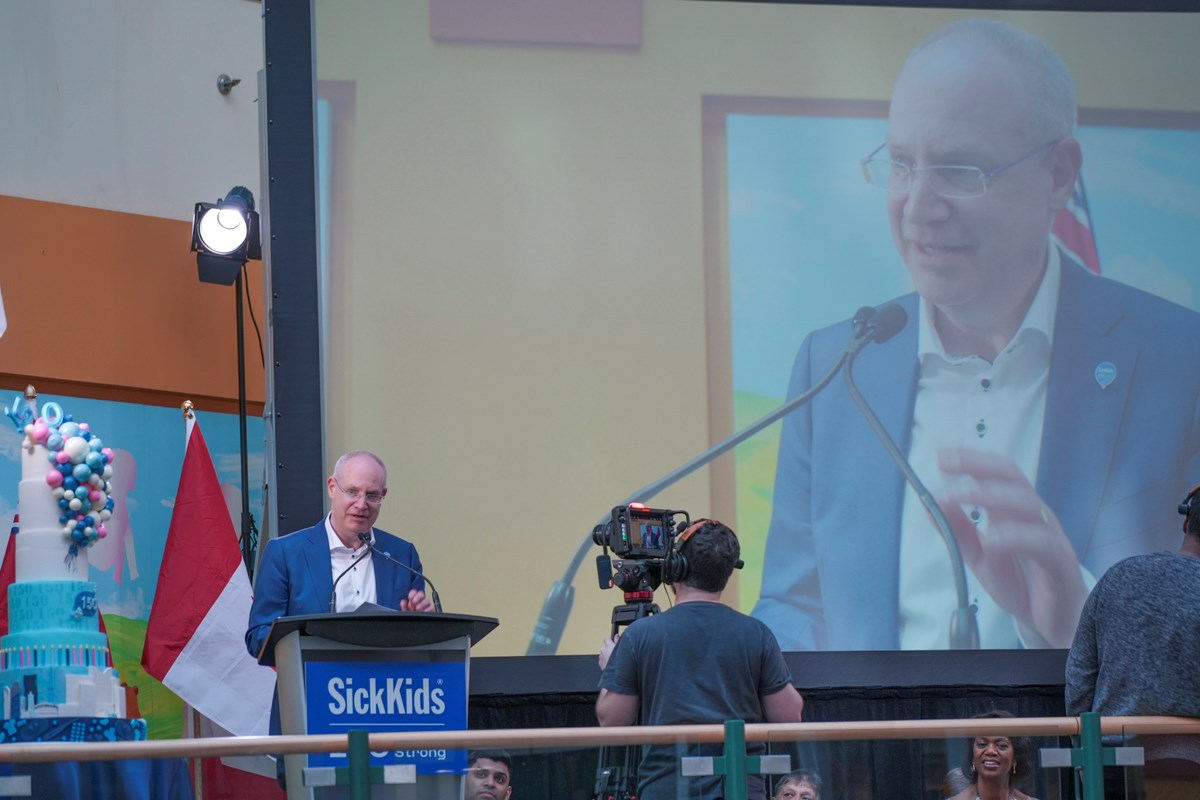 Dr. Ronald Cohn delivering a speech at a podium on the stage in the Atrium. Next to him is a multilayer birthday cake.
